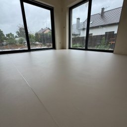 Technodecor Yevhen Syvodiedov - Jasna, gładka posadzka w nowym wnętrzu z dużymi oknami, przez które widać dom i ogród. Minimalistyczny design, świeżo wylana podłoga, widoczne łączenia.