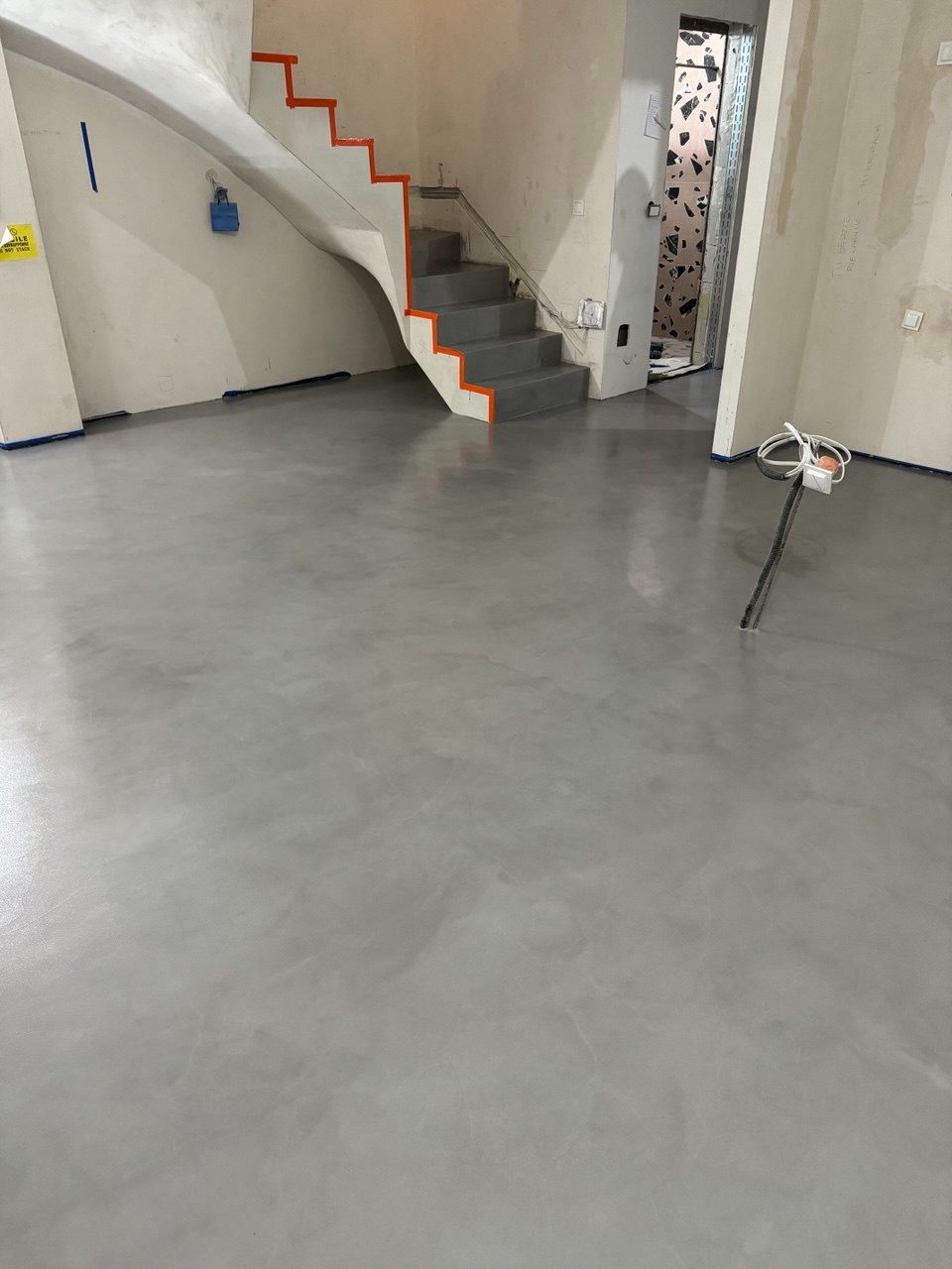 Szara, gładka posadzka w jasnym wnętrzu z białymi schodami o pomarańczowych krawędziach. Widoczne niedokończone prace wykończeniowe i wystające kable.