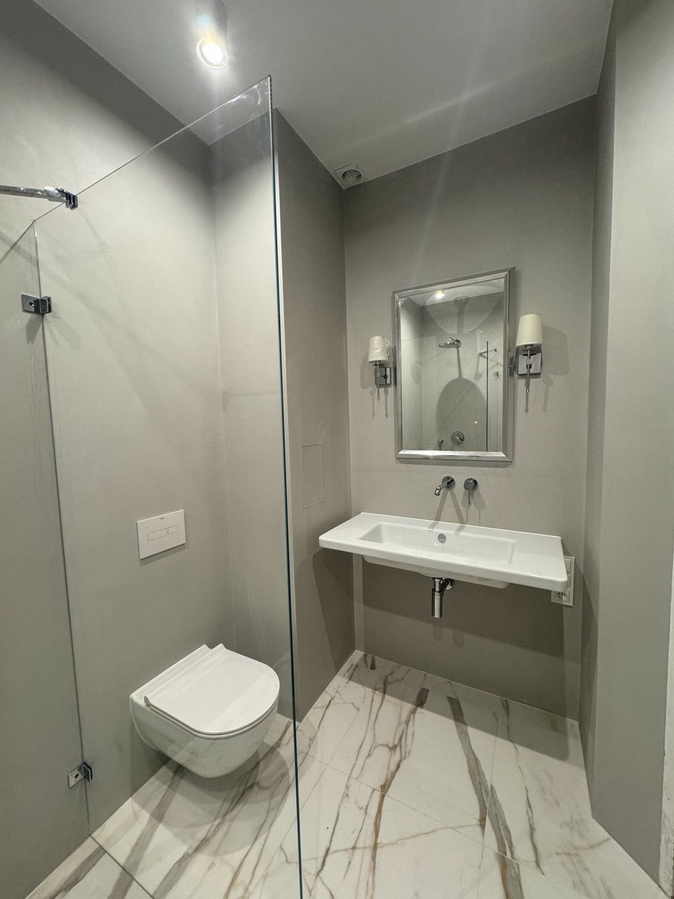 Szara łazienka z kabiną typu walk-in, umywalką i toaletą podwieszaną. Marmurowe płytki podłogowe z brązowymi żyłami. Nowoczesne oświetlenie.