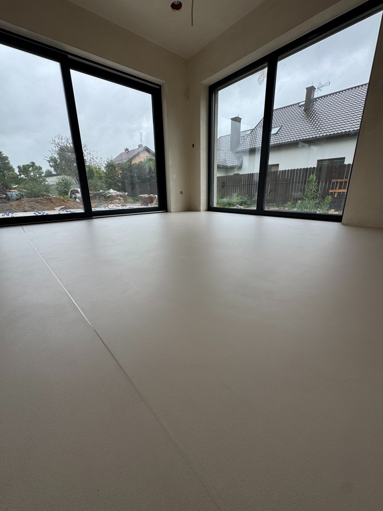 Jasna, gładka posadzka w nowym wnętrzu z dużymi oknami, przez które widać dom i ogród. Minimalistyczny design, świeżo wylana podłoga, widoczne łączenia.