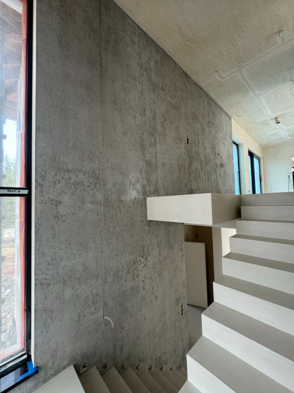 Ściana z szarą, dekoracyjną powłoką imitującą beton, sąsiadująca z białymi schodami i oknem. Widoczne niedoskonałości faktury i surowy charakter wykończenia.