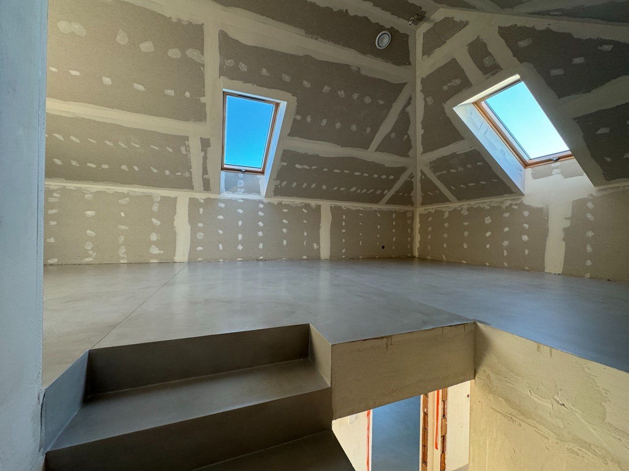 Szara, gładka posadzka w surowym wnętrzu poddasza ze skosami i oknami dachowymi. Ściany przygotowane do malowania. Widoczne schody z posadzką.