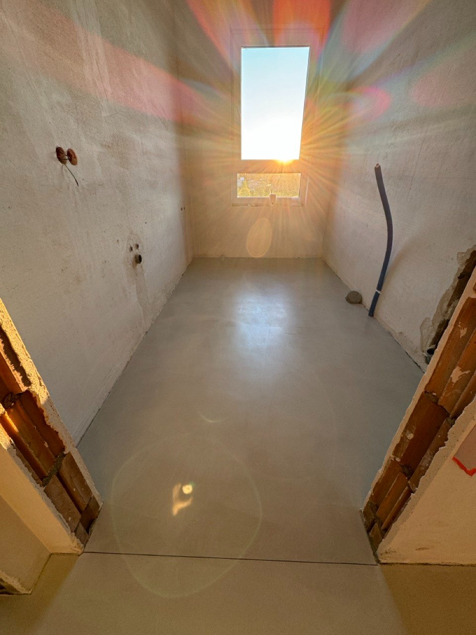 Jasna, gładka posadzka w surowym wnętrzu z oknem dachowym, przez które wpada silne światło słoneczne. Widoczne fragmenty ścian w trakcie wykończenia i instalacja.