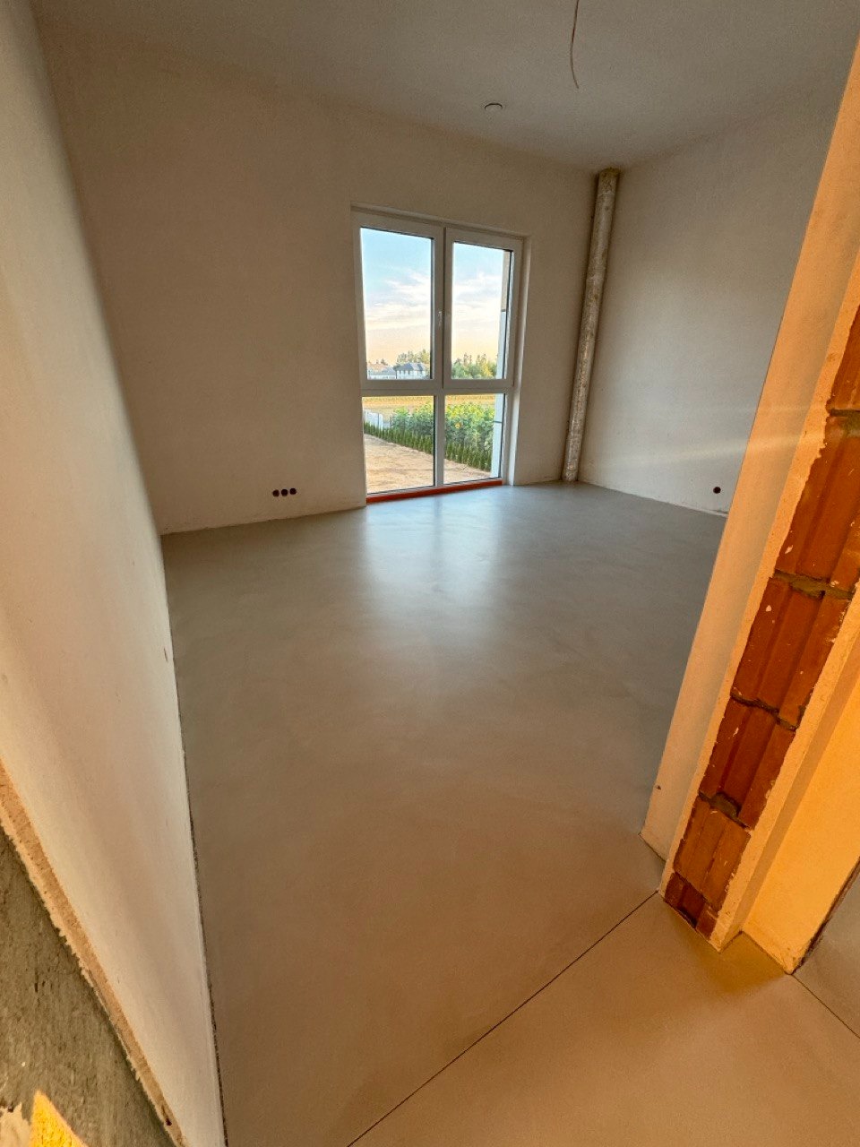 Jasnoszara, gładka posadzka w pustym pokoju z oknem i widokiem na zieleń. Minimalistyczne wnętrze w surowym stylu, z widocznymi elementami konstrukcyjnymi.