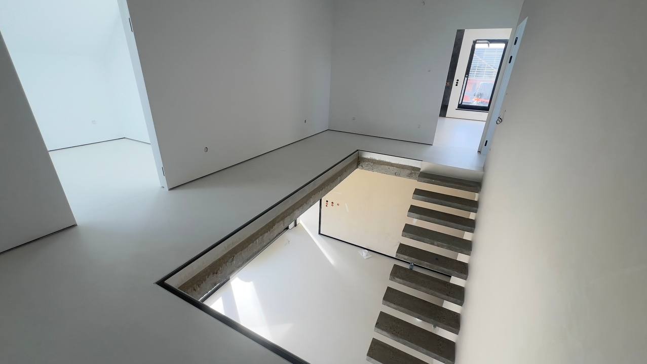 Minimalistyczne wnętrze z posadzką z mikrocementu i schodami w dół. Jasne, jednolite wykończenie podłogi i ścian. Widok z góry na schody.