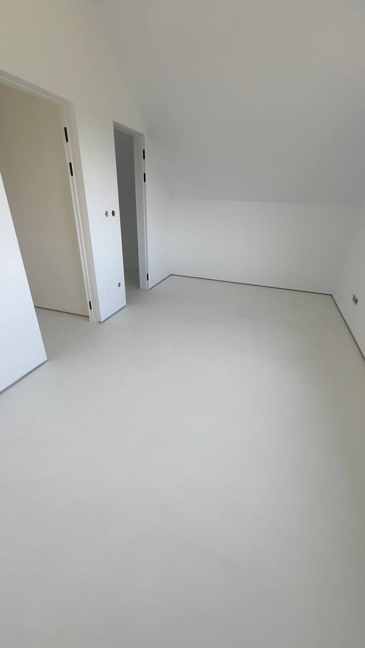 Jasne, minimalistyczne wnętrze poddasza z gładką, jednolitą posadzką. Dwa białe otwory drzwiowe i skosy sufitu podkreślają nowoczesny charakter wykończenia.