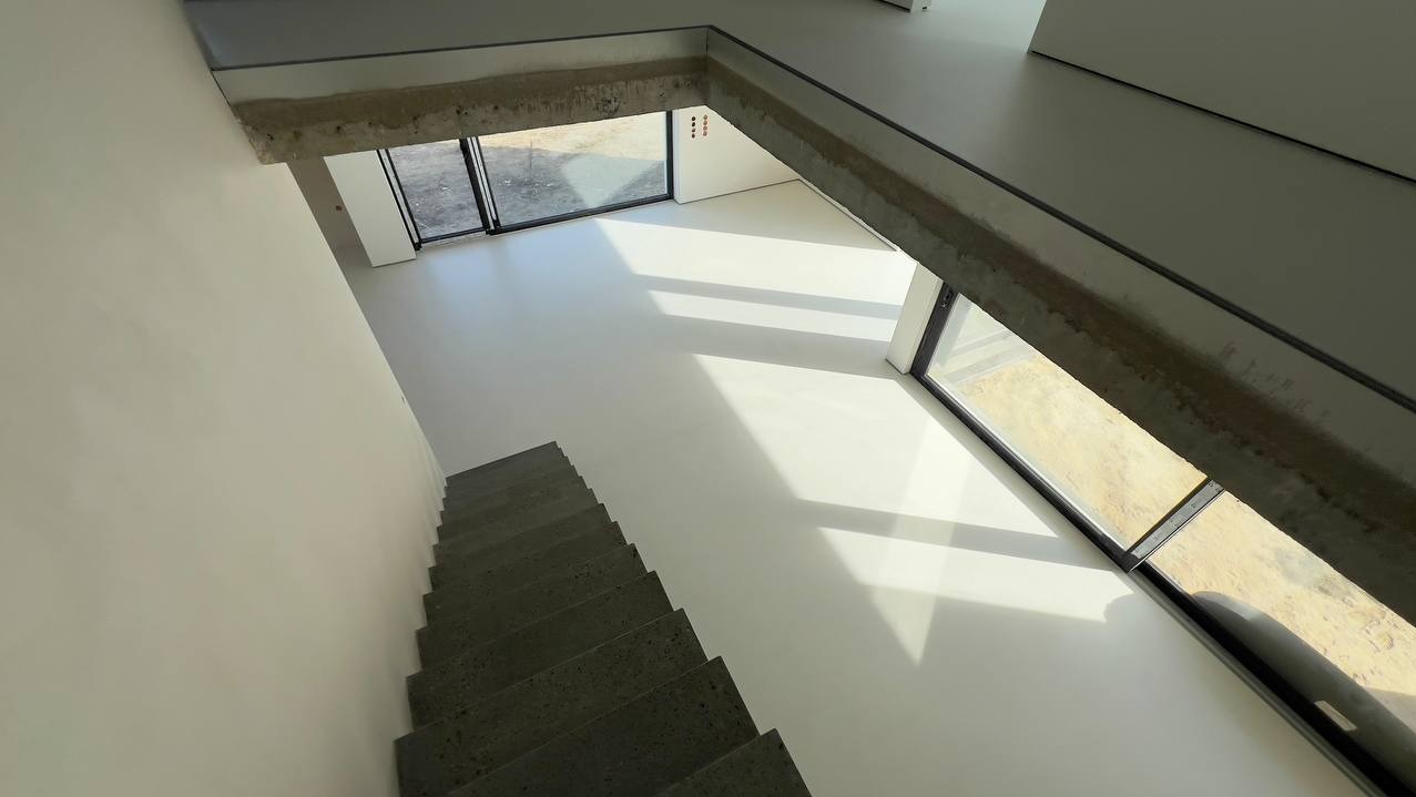 Jasna, gładka posadzka w nowoczesnym wnętrzu z betonowymi schodami i dużymi oknami. Widok z góry, podkreślający minimalizm i przestronność.