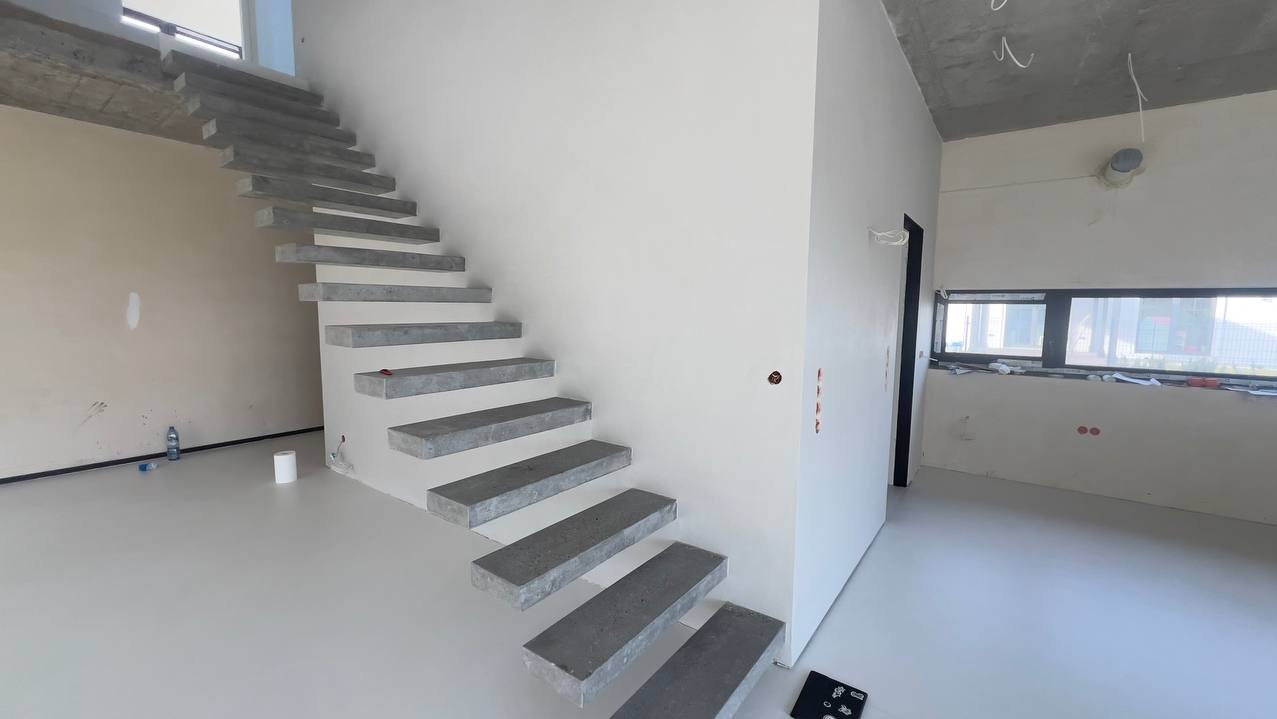 Minimalistyczne schody z szarego betonu prowadzą na górę w jasnym, nowoczesnym wnętrzu. Gładka, jasna posadzka i białe ściany podkreślają surowy charakter wykończenia.