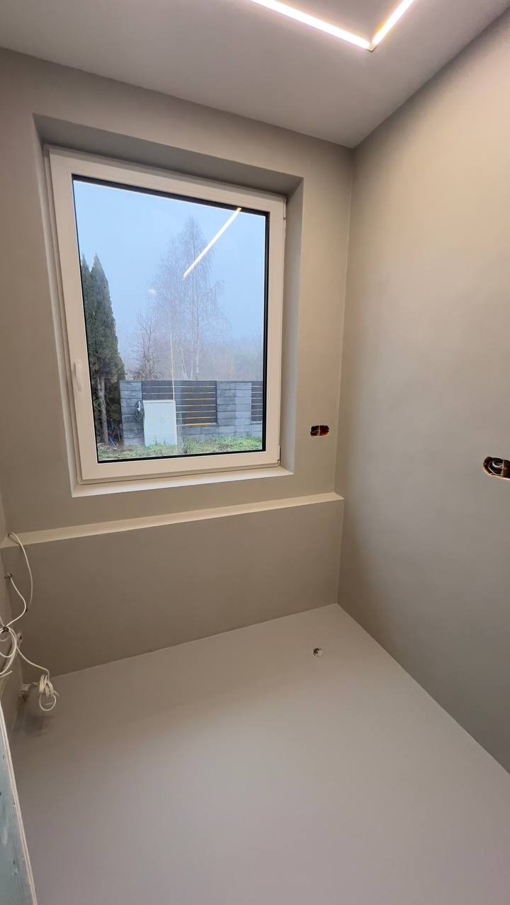 Jasne wnętrze po wykończeniu ścian i podłogi jednolitą, gładką powierzchnią. Widoczne okno z widokiem na zewnątrz i minimalistyczne oświetlenie sufitowe.