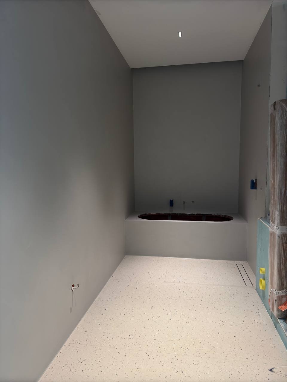 Biała posadzka z drobnymi plamkami w łazience w Warszawie. Minimalistyczne wnętrze z wanną w zabudowie i szarą ścianą. Widoczny odpływ liniowy.