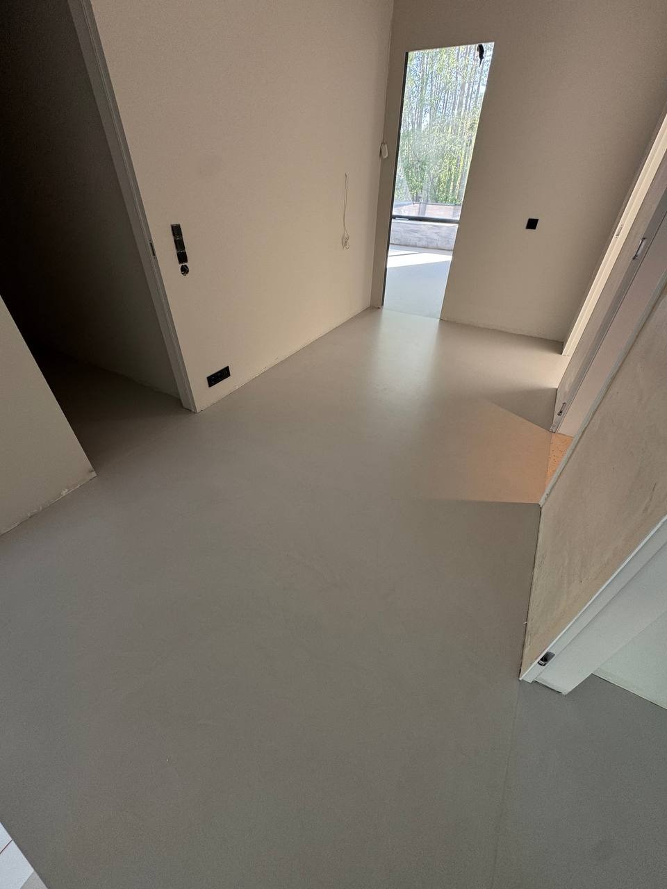 Jasnoszara, gładka posadzka z mikrocementu w nowoczesnym wnętrzu. Widoczne ściany w odcieniach bieli i fragment okna z widokiem na zieleń. Minimalistyczny design.