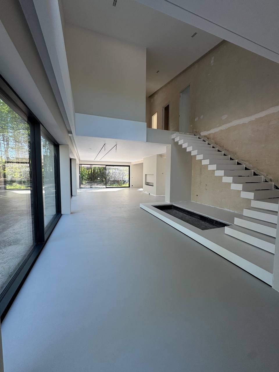 Jasnoszara posadzka w przestronnym wnętrzu z dużymi oknami i białymi schodami. Minimalistyczny design z elementami surowego betonu na ścianie przy schodach.