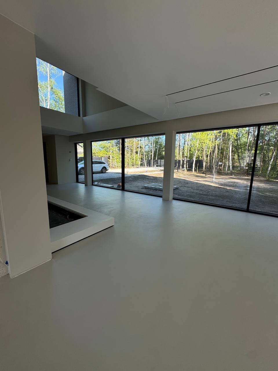 Jasnoszara, gładka posadzka w nowoczesnym salonie z dużymi oknami. Minimalistyczny design, widok na zieleń za oknem. Nowoczesny kominek wkomponowany w podłogę.