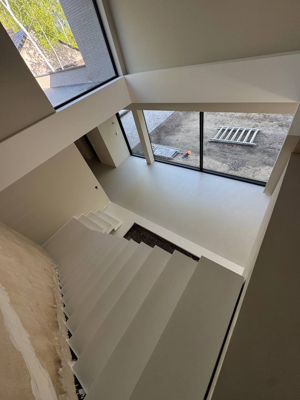 Jasne wnętrze domu po remoncie: schody i podłoga pokryte jednolitą, gładką powierzchnią. Widok z góry, duże okna z widokiem na zewnątrz, drabina oparta o ścianę.