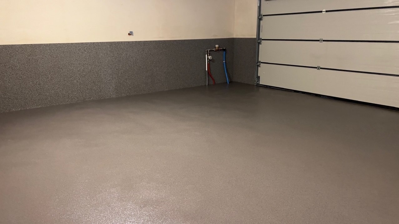 Szara, gładka posadzka w garażu z częściowo otwartą białą bramą segmentową. Widoczne przyłącza wodne na ścianie z szarym wykończeniem do połowy wysokości.
