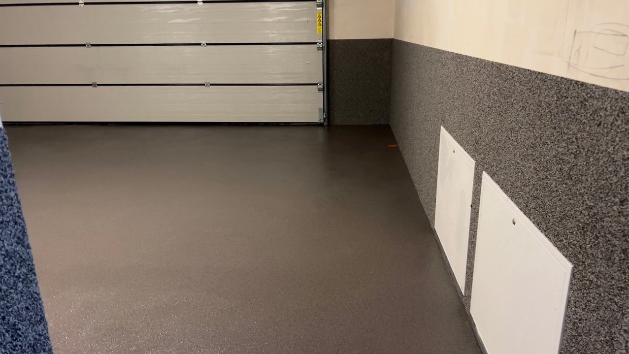 Gładka, brązowa posadzka w garażu z szarą ścianą wykończoną dekoracyjną powłoką. Widoczne białe klapy rewizyjne i fragment otwartej bramy garażowej.