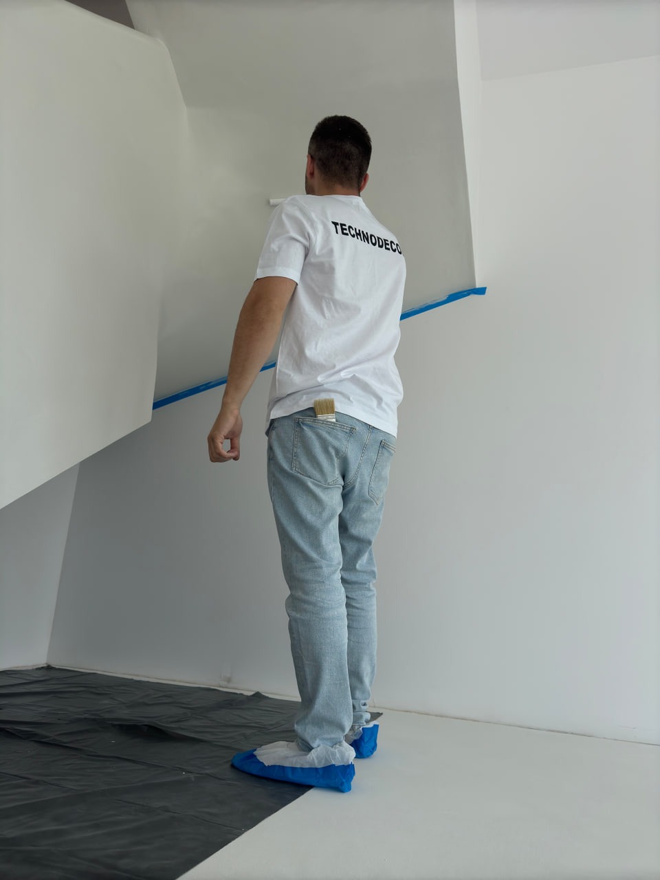 Pracownik w trakcie aplikacji białej powłoki na schodach, zabezpieczony folią i ochraniaczami na obuwiu. Pędzel w kieszeni spodni. Widok z tyłu, logo na koszulce.