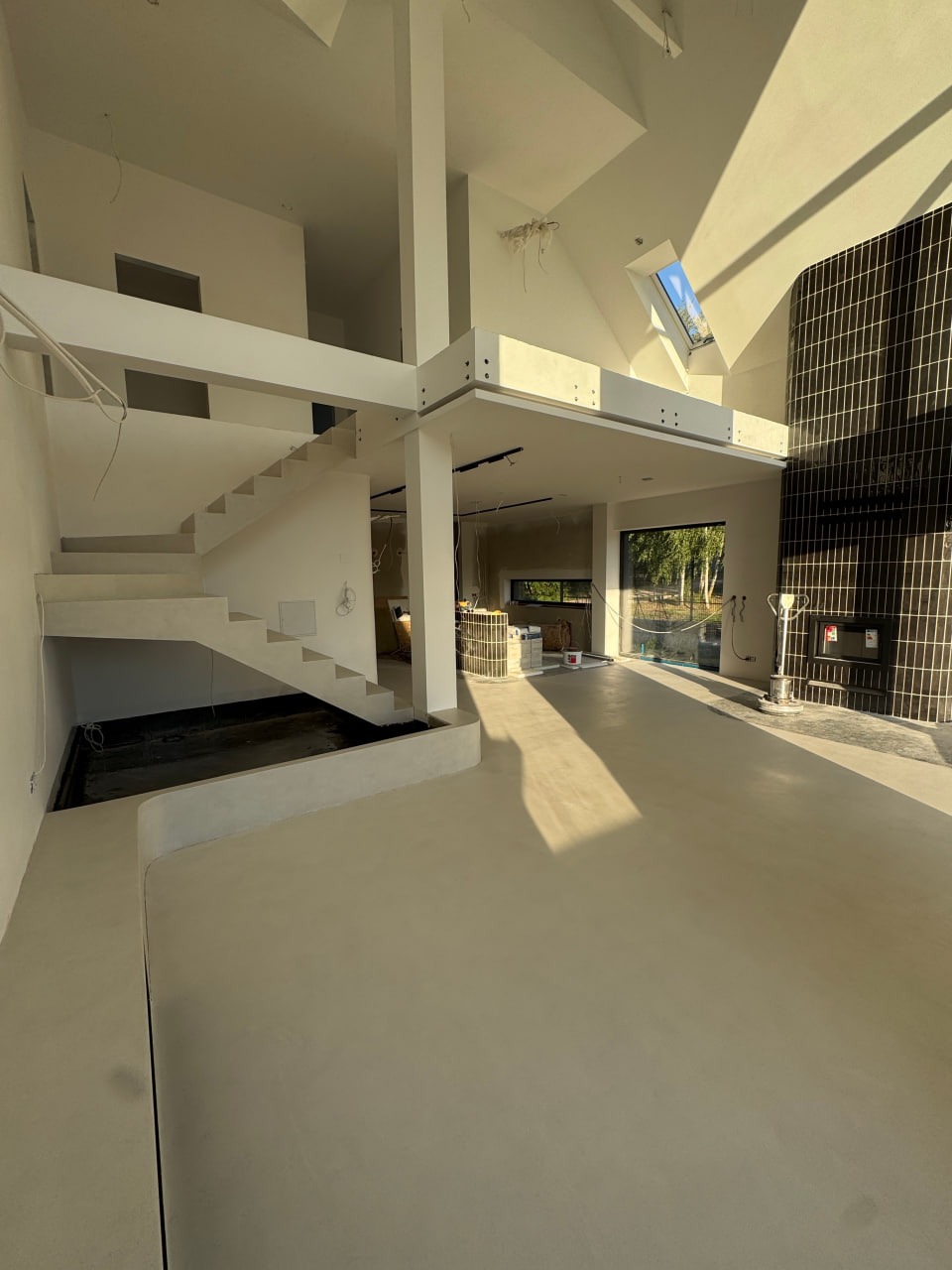 Jasna, przestronna przestrzeń z gładką, jednolitą posadzką. Schody z minimalistyczną balustradą, duże okno i czarna ściana z płytek dodają nowoczesnego charakteru.