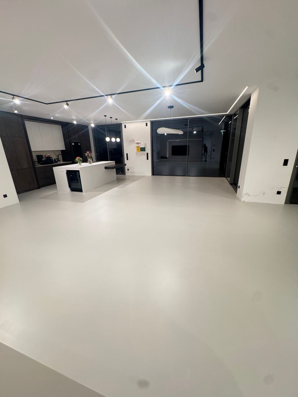 Jasna, nowoczesna przestrzeń z gładką, jednolitą posadzką w odcieniach szarości. Widoczna kuchnia z wyspą i przeszklone drzwi przesuwne. Minimalistyczne oświetlenie.