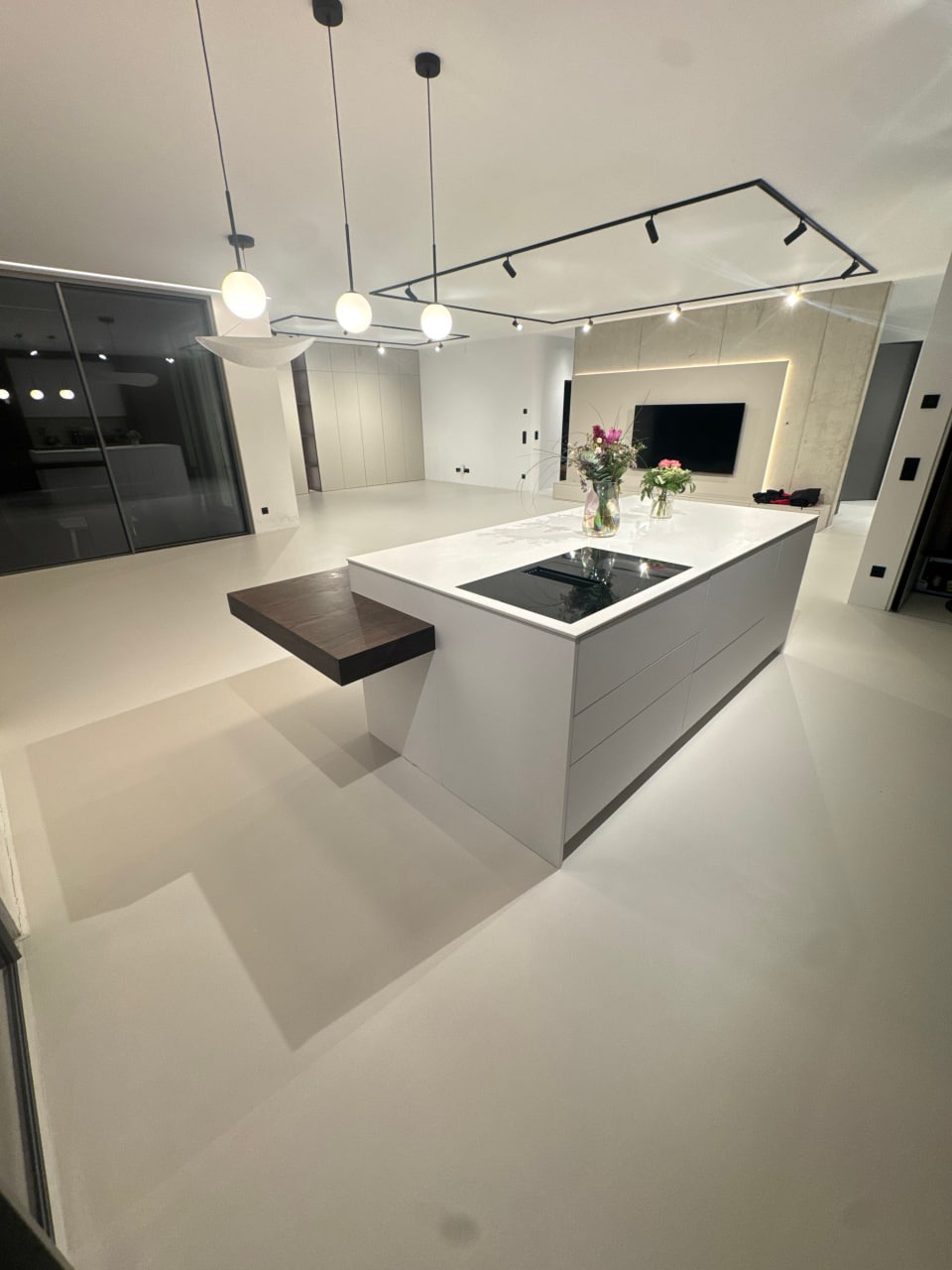 Minimalistyczne wnętrze z jasną posadzką, wyspą kuchenną z blatem imitującym marmur i nowoczesnym oświetleniem. Gładka powierzchnia podłogi odbija światło.
