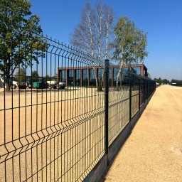 PBPW Paulina Posak-Sołtysiak - Długi, czarny panelowy system ogrodzeniowy z betonową podmurówką, ciągnący się wzdłuż żwirowej drogi, z budynkiem w tle i maszynami budowlanymi.