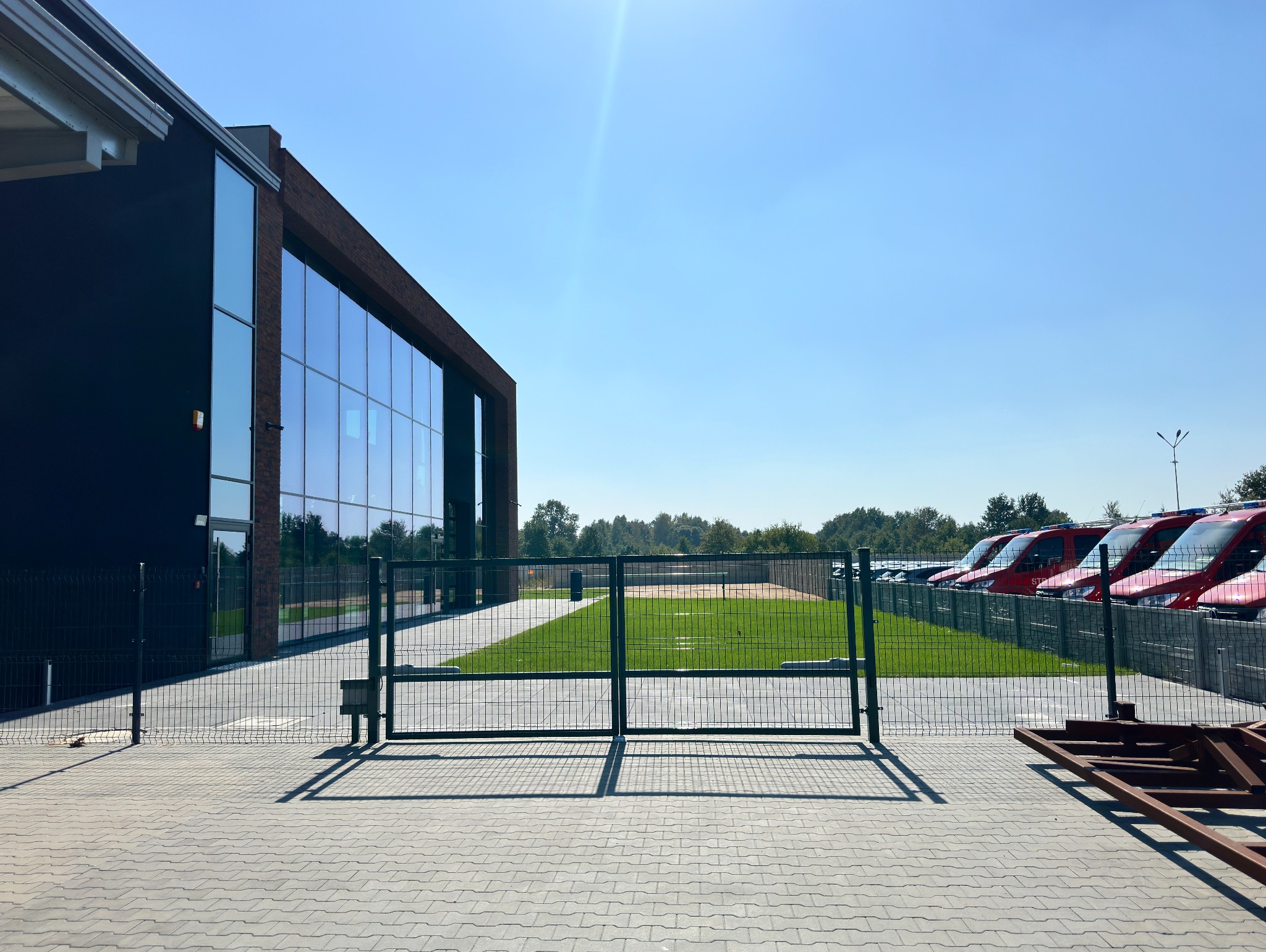 Nowoczesna brama wjazdowa z metalową siatką na tle budynku z przeszkloną fasadą i rzędem czerwonych samochodów. Jasne, słoneczne oświetlenie.