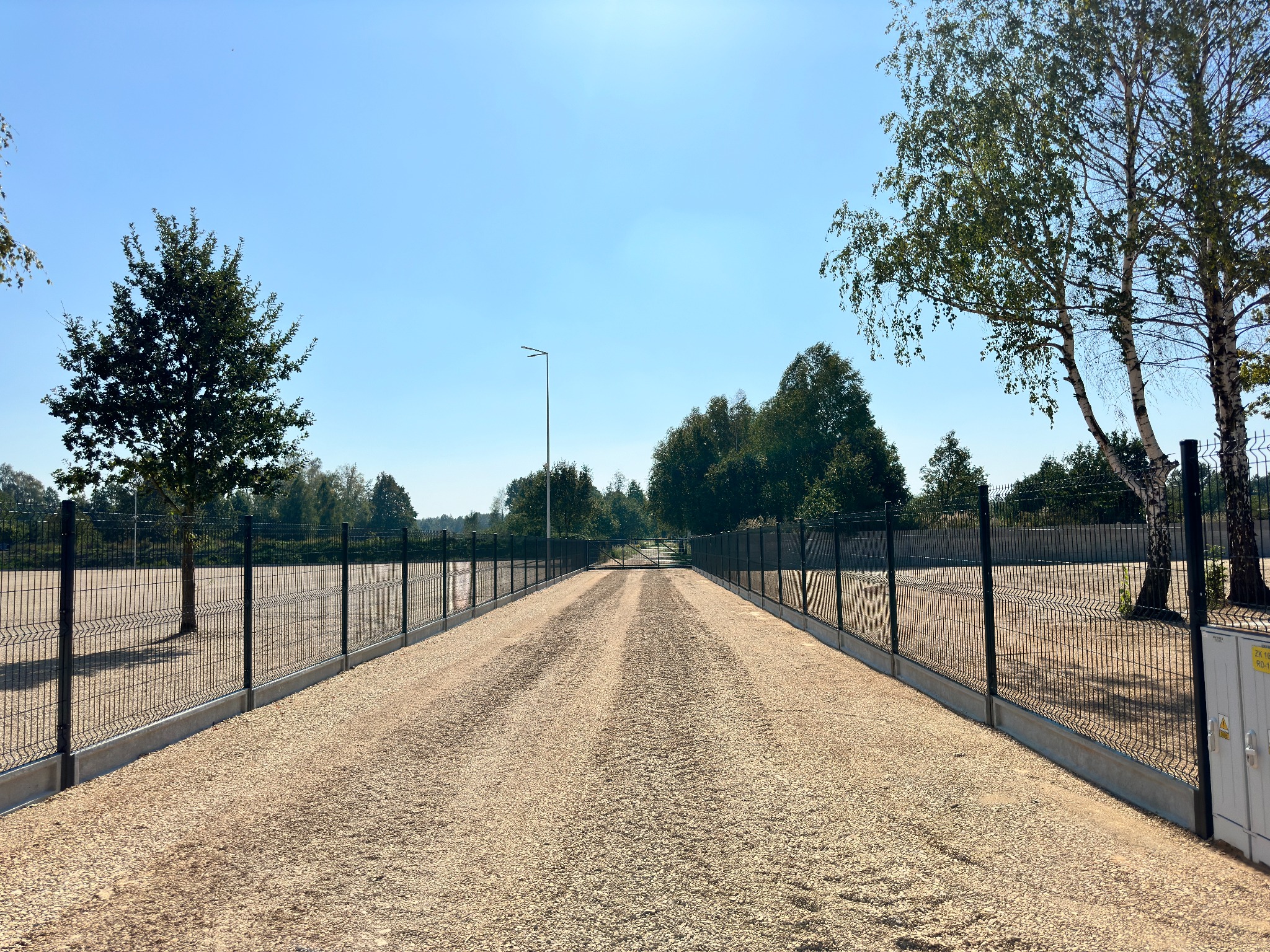 Prosta droga wysypana żwirem, po bokach ogrodzenie panelowe w kolorze grafitowym, w tle drzewa i błękitne niebo. Perspektywa centralna.