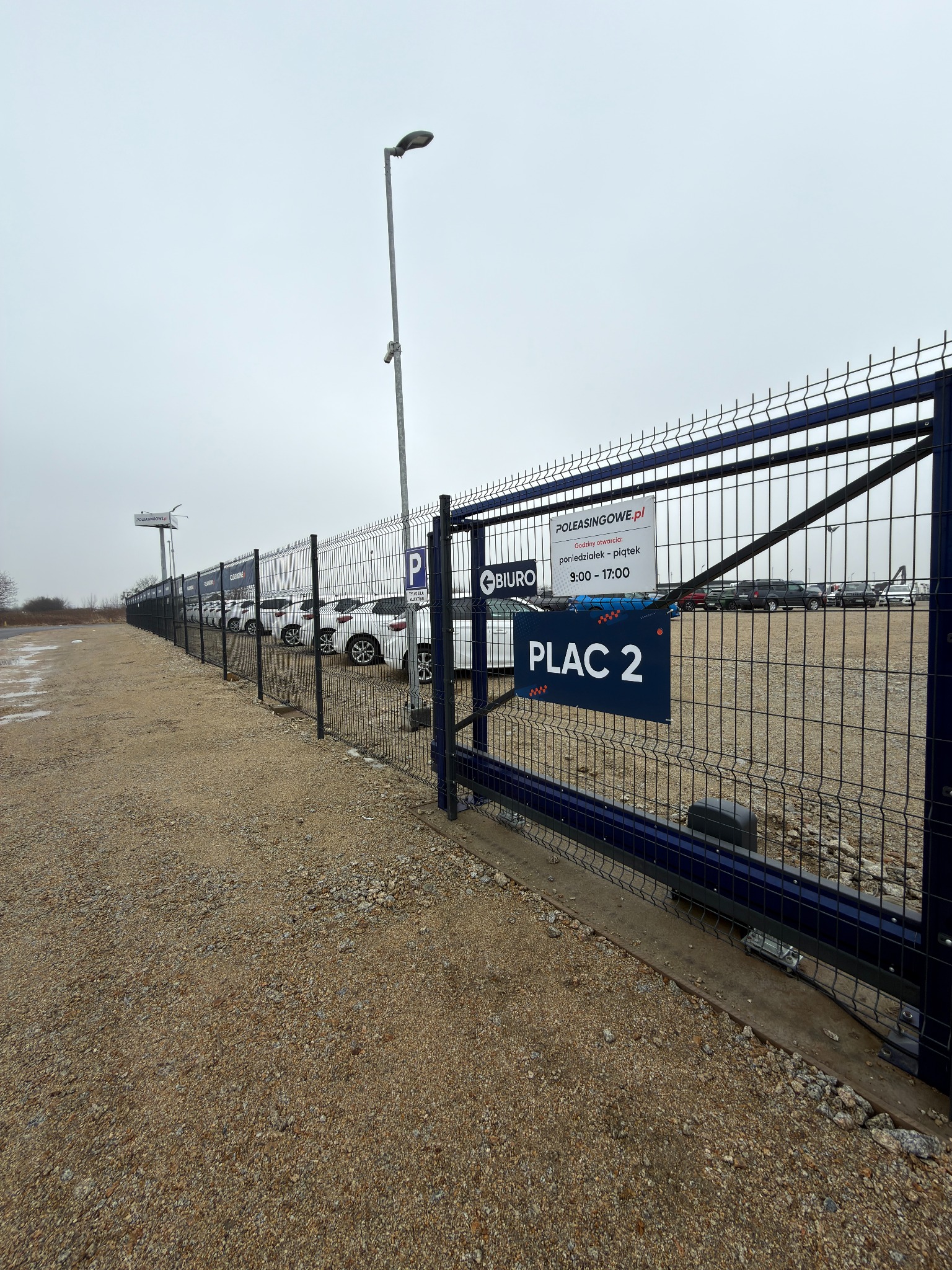 Brama wjazdowa z siatki z tabliczką 'Plac 2' na tle parkingu samochodowego. Widoczny mechanizm otwierania bramy i słup oświetleniowy. Pochmurne niebo.