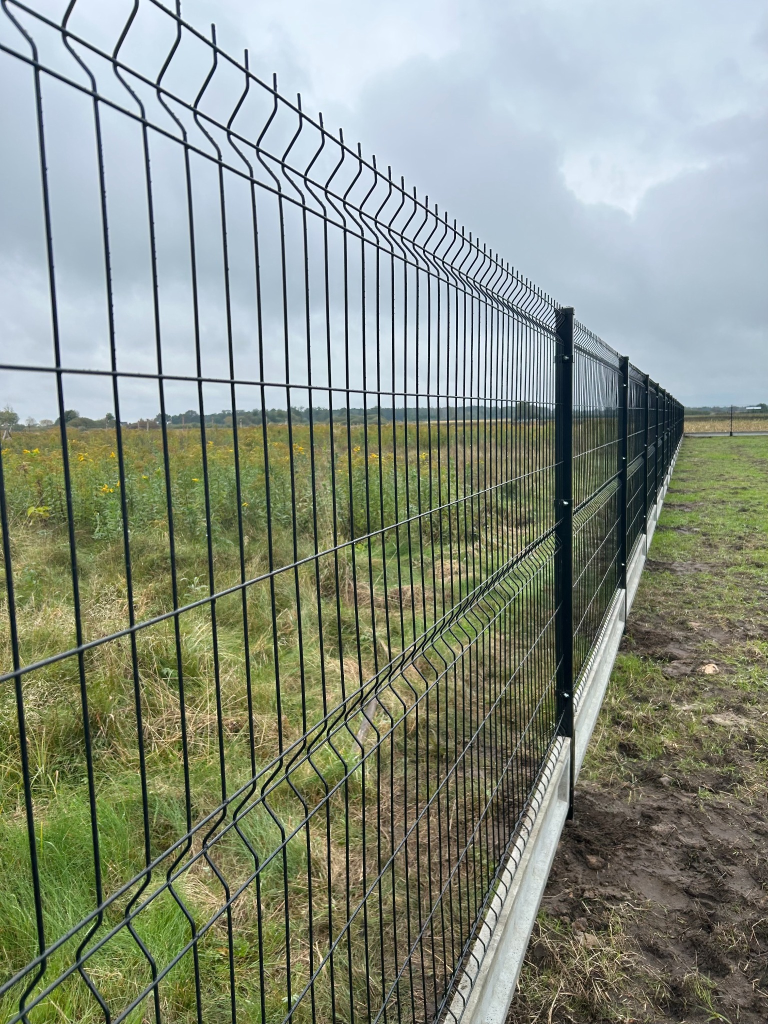 Długi, czarny panel ogrodzeniowy z podmurówką betonową, ciągnący się wzdłuż zielonego pola pod pochmurnym niebem. Widok z perspektywy, ukazujący głębię ogrodzenia.