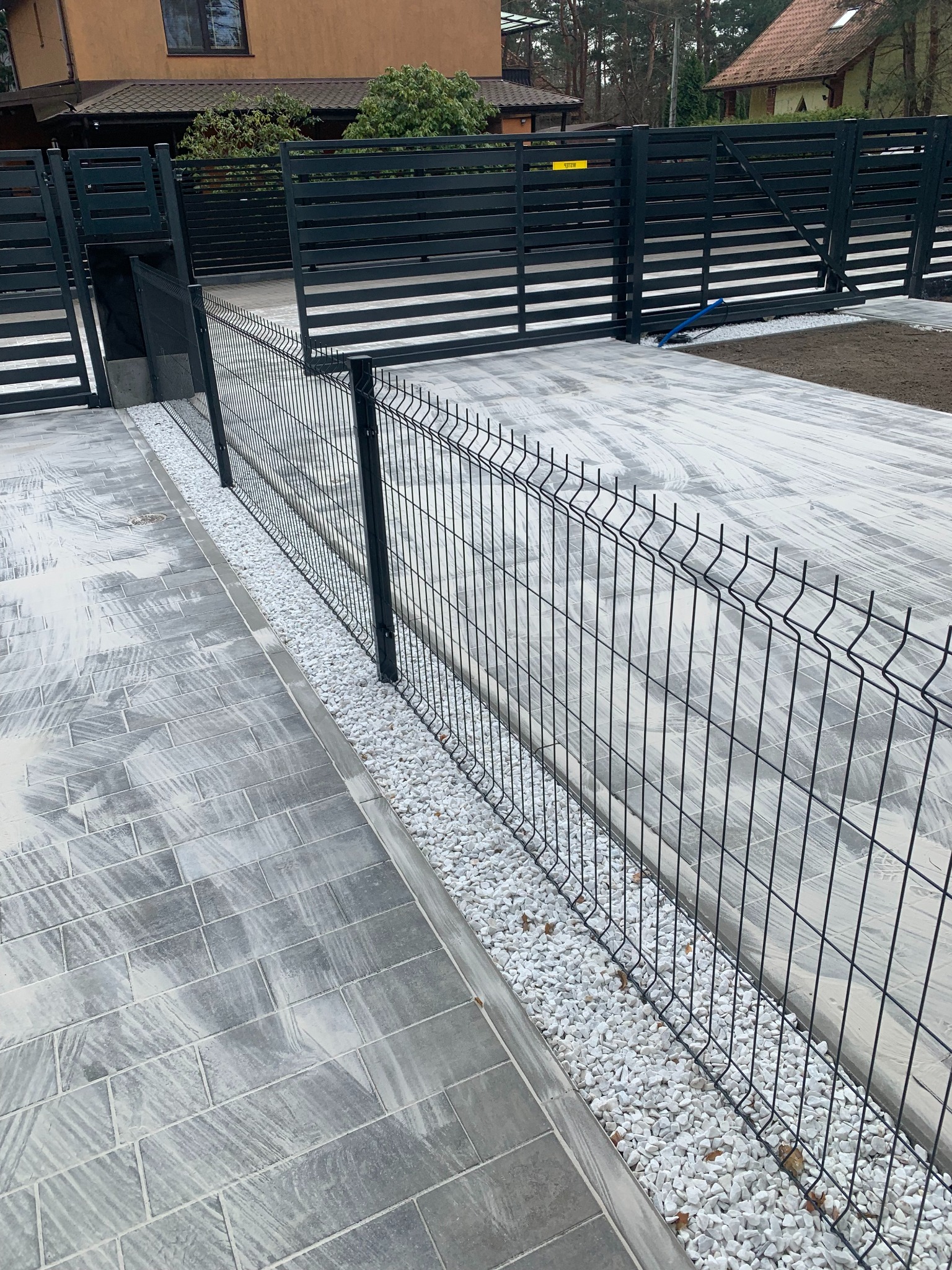 Nowoczesne ogrodzenie panelowe z bramą przesuwną, wykończone białym kamieniem. Szara kostka brukowa i dom w tle. Ujęcie z niskiej perspektywy.