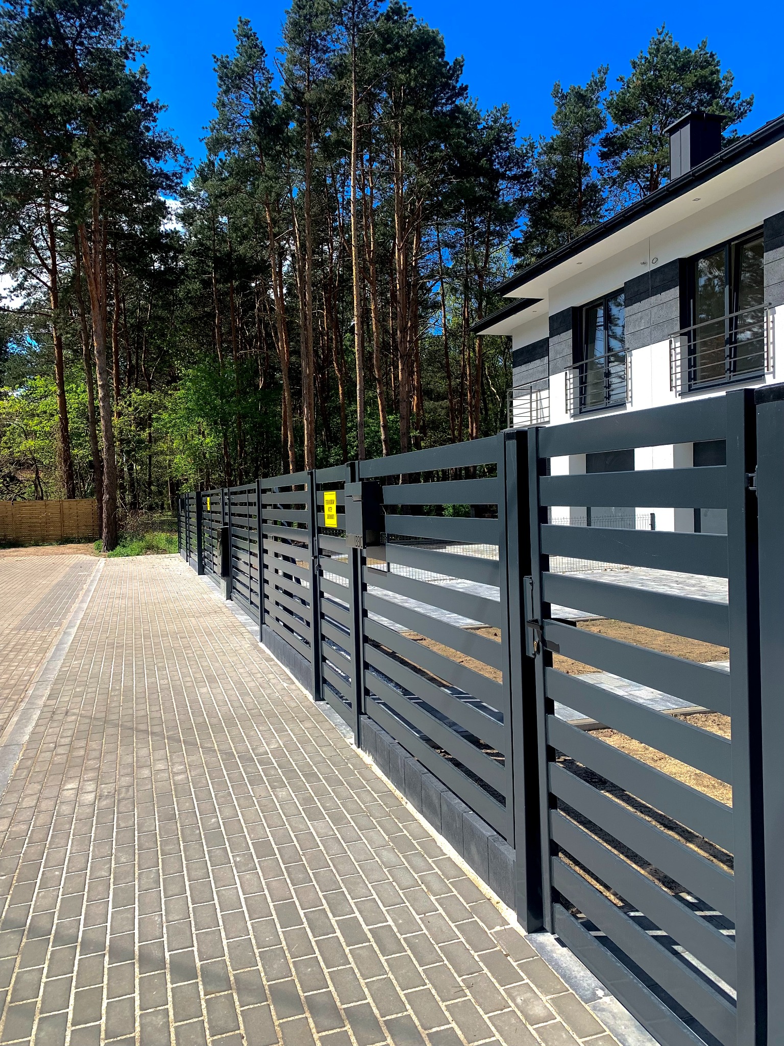 Nowoczesne, poziome ogrodzenie metalowe w kolorze grafitowym, z widocznym fragmentem domu w tle. Ułożona kostka brukowa prowadzi wzdłuż ogrodzenia.