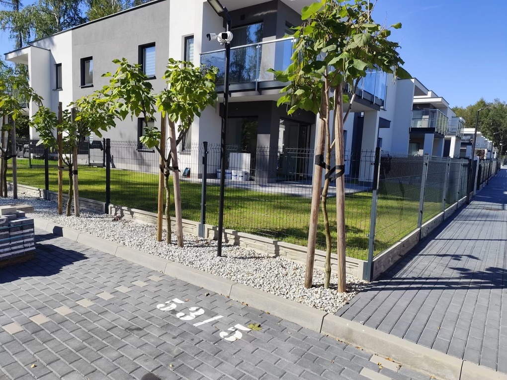 Nowoczesne osiedle z niskim ogrodzeniem panelowym w kolorze grafitowym, oddzielającym budynki od chodnika z kostki. Młode drzewka posadzone wzdłuż ogrodzenia.