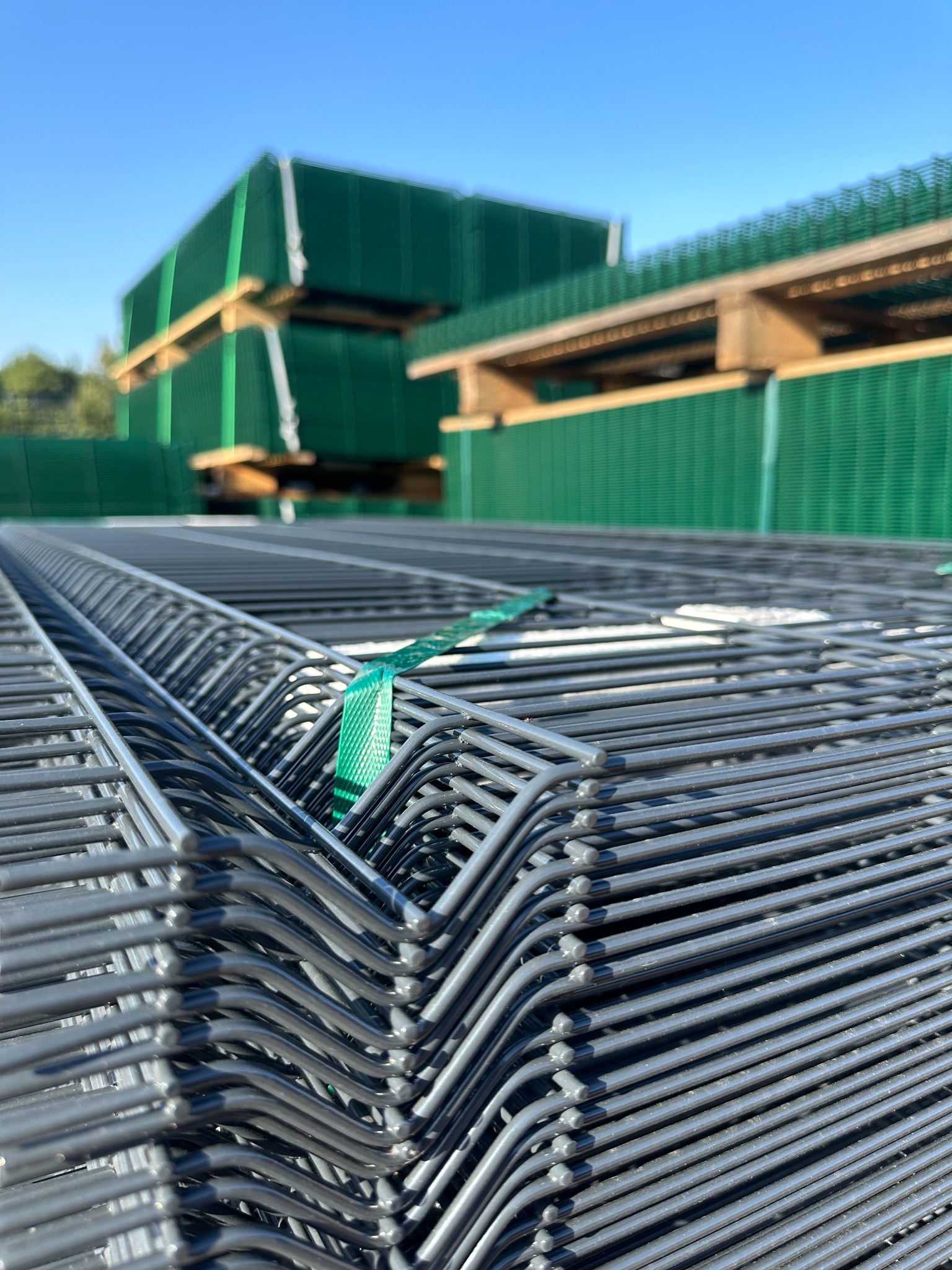 Stos paneli ogrodzeniowych w kolorze szarym na pierwszym planie, w tle zielone panele na paletach pod błękitnym niebem. Zabezpieczone zieloną taśmą.