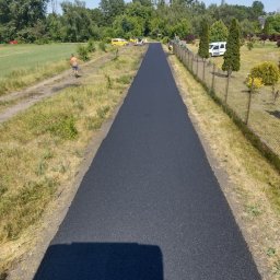 Nowa, asfaltowa droga dojazdowa na tle zieleni i błękitnego nieba. Widoczne maszyny budowlane i pracownik. Świeżo położony asfalt, równa powierzchnia.