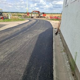 Świeżo ułożona, zakrzywiona nawierzchnia asfaltowa z koparką w tle i turbiną wiatrową w oddali, przylegająca do ściany budynku z widocznym korytkiem odwadniającym.