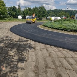 Walec drogowy ubija świeżo wylany asfalt na krętej drodze. W tle zbiorniki gazu i pracownik w kamizelce odblaskowej. Słoneczny dzień, błękitne niebo z chmurami.