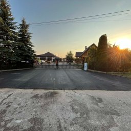 Nowa, gładka nawierzchnia asfaltowa przed posesją z bramą wjazdową. W tle budynki i drzewa iglaste, a w oddali zachodzące słońce.