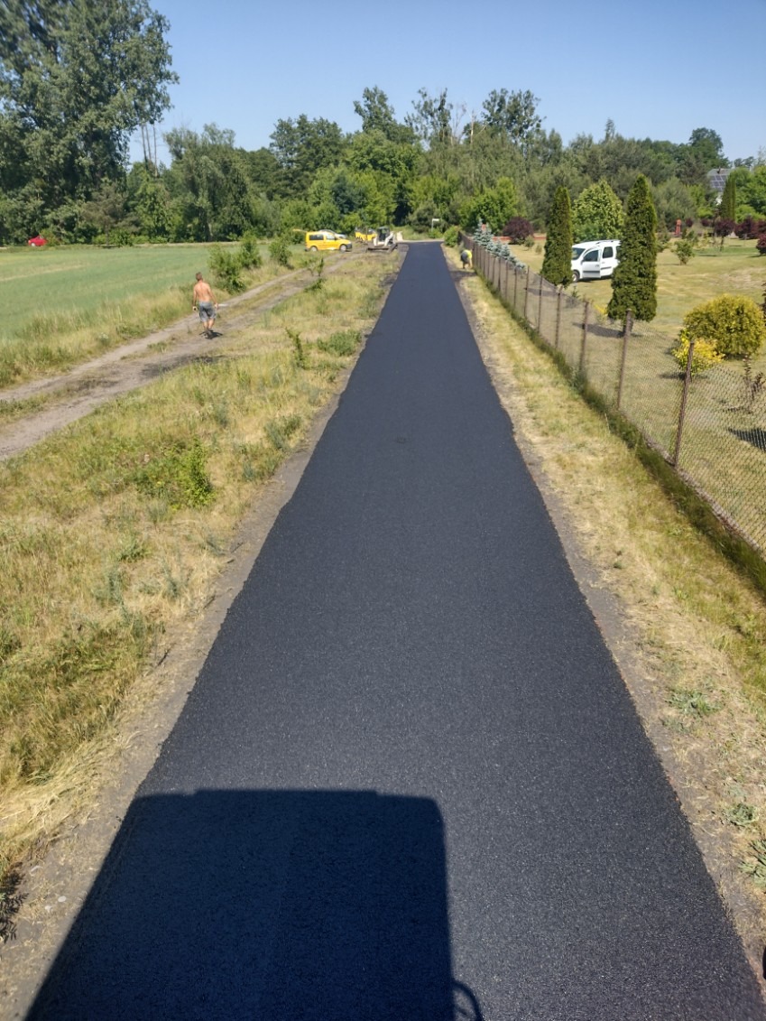 Nowa, asfaltowa droga dojazdowa na tle zieleni i błękitnego nieba. Widoczne maszyny budowlane i pracownik. Świeżo położony asfalt, równa powierzchnia.