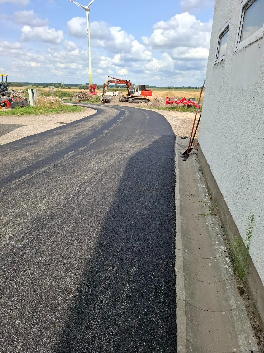Świeżo ułożona, zakrzywiona nawierzchnia asfaltowa z koparką w tle i turbiną wiatrową w oddali, przylegająca do ściany budynku z widocznym korytkiem odwadniającym.