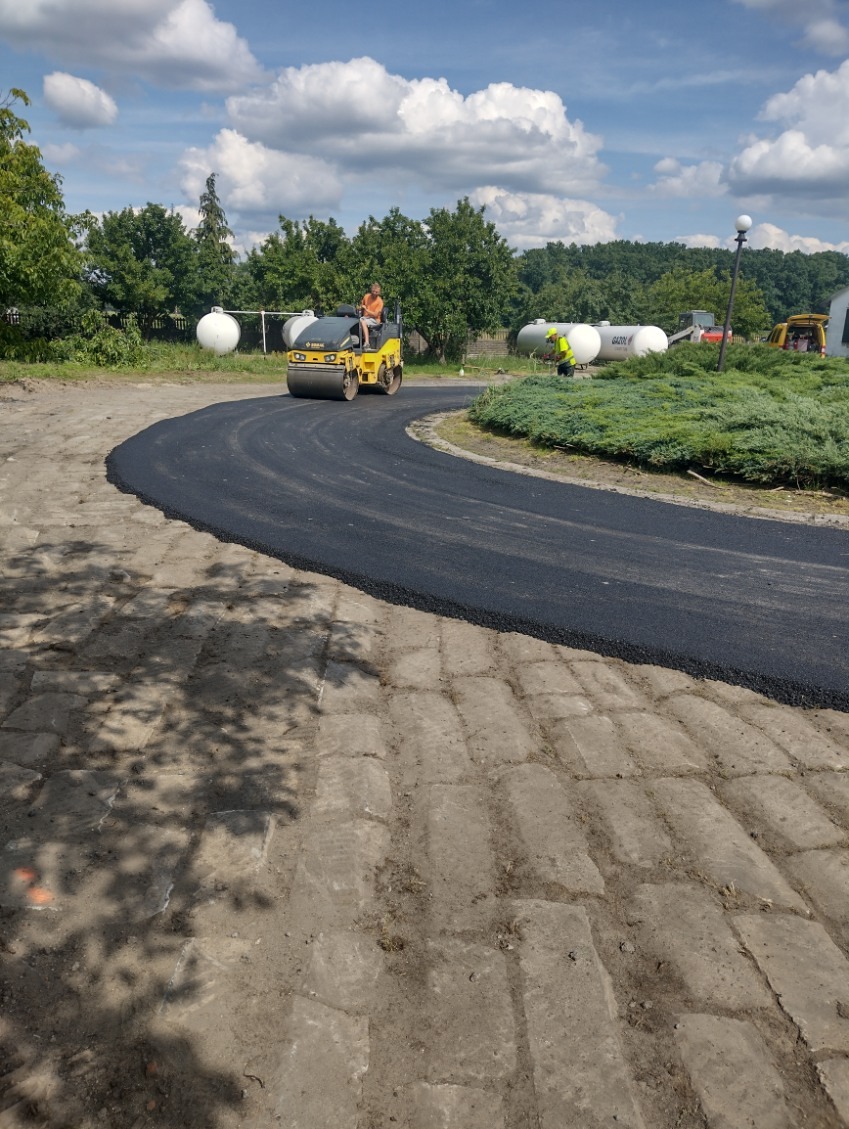 Walec drogowy ubija świeżo wylany asfalt na krętej drodze. W tle zbiorniki gazu i pracownik w kamizelce odblaskowej. Słoneczny dzień, błękitne niebo z chmurami.