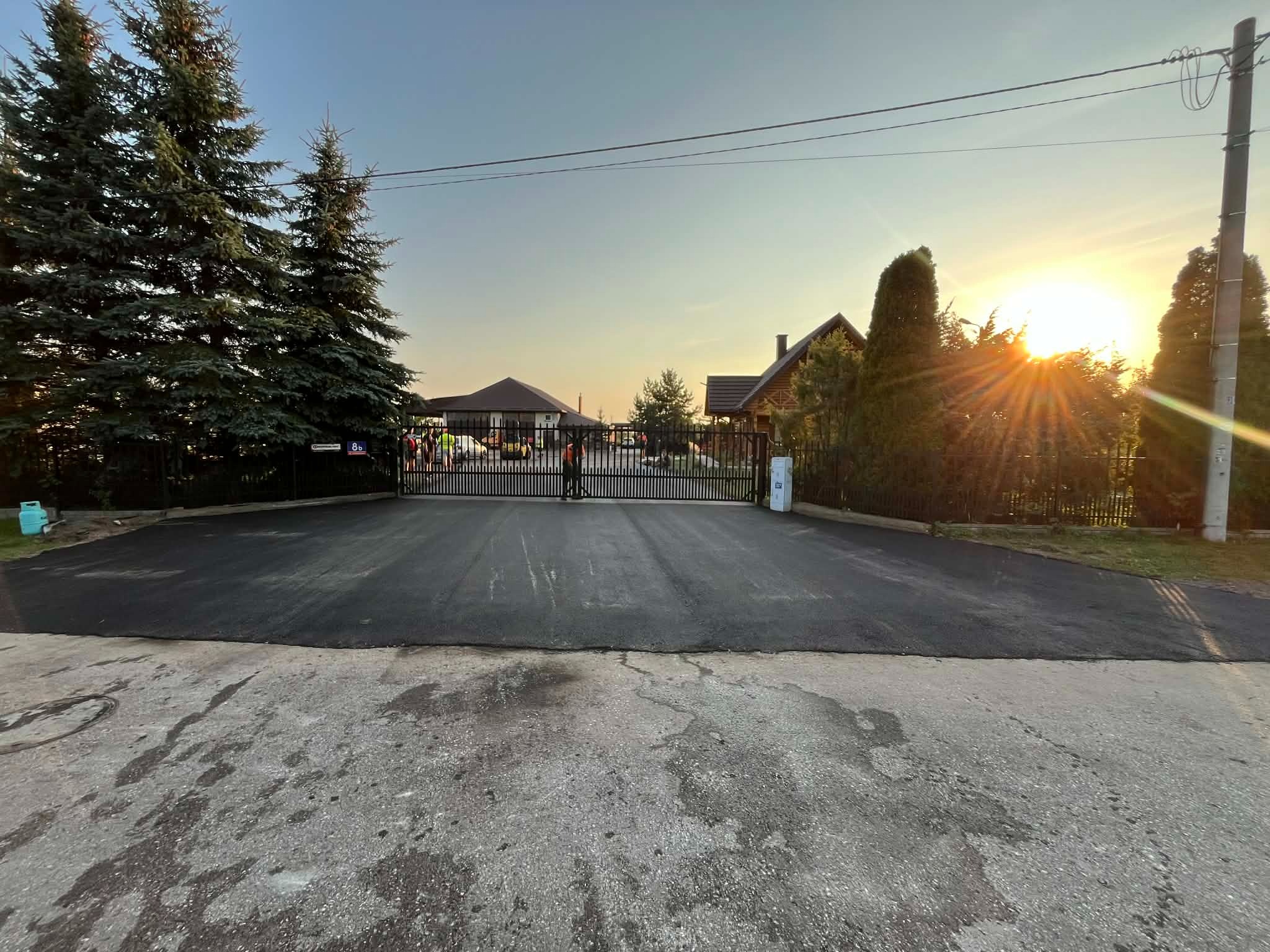 Nowa, gładka nawierzchnia asfaltowa przed posesją z bramą wjazdową. W tle budynki i drzewa iglaste, a w oddali zachodzące słońce.
