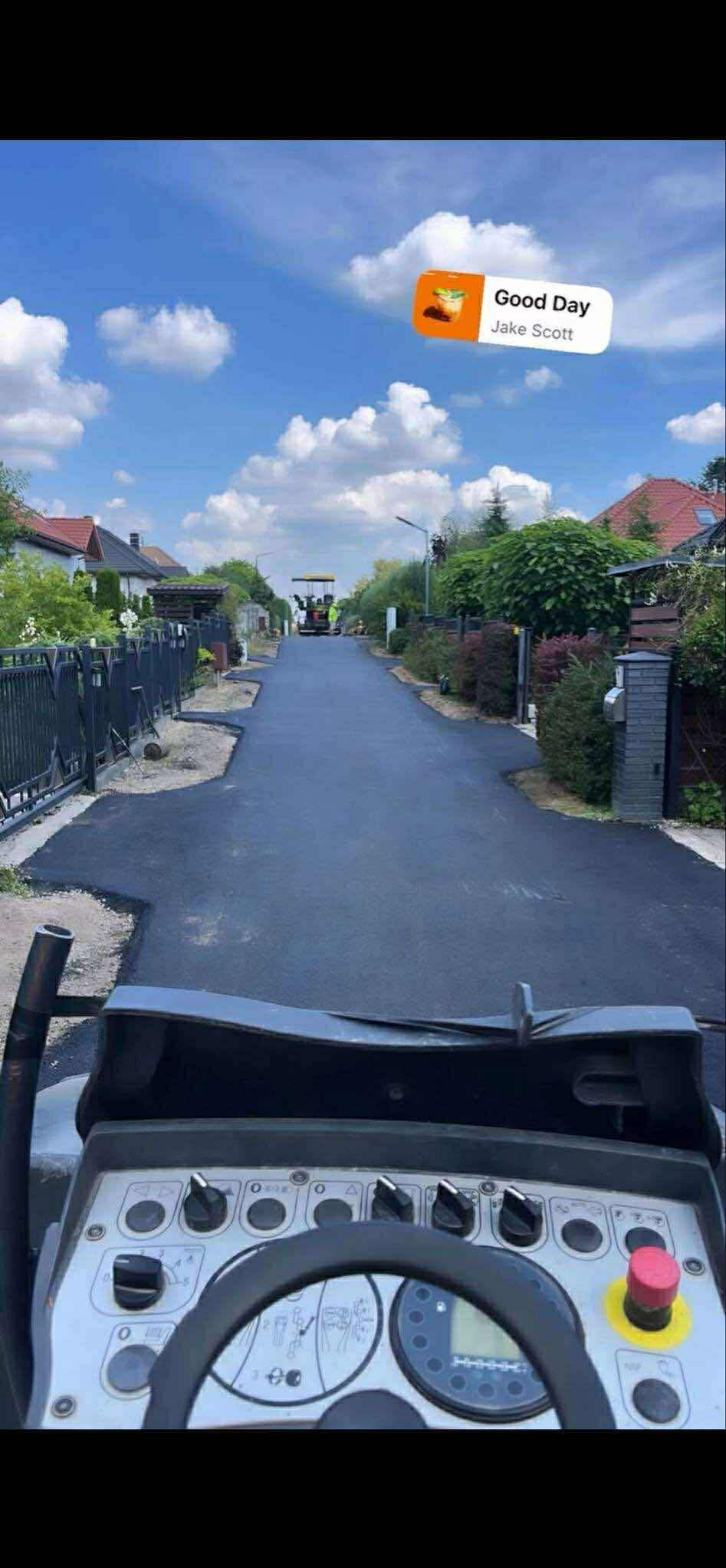 Widok z kabiny maszyny na świeżo wyasfaltowaną drogę w otoczeniu domów i zieleni, pod błękitnym niebem z chmurami. W tle widoczna ekipa pracująca przy rozkładaniu asfaltu.