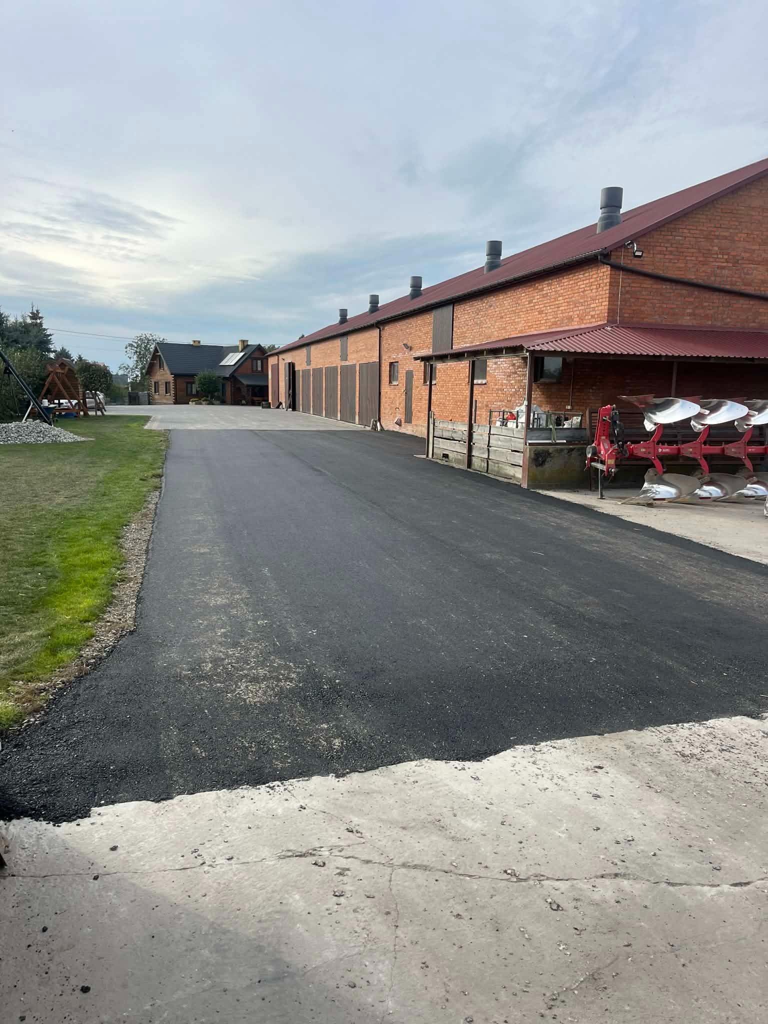 Nowa, asfaltowa droga dojazdowa na posesji z budynkami z czerwonej cegły w tle. Po prawej stronie widoczny sprzęt rolniczy.