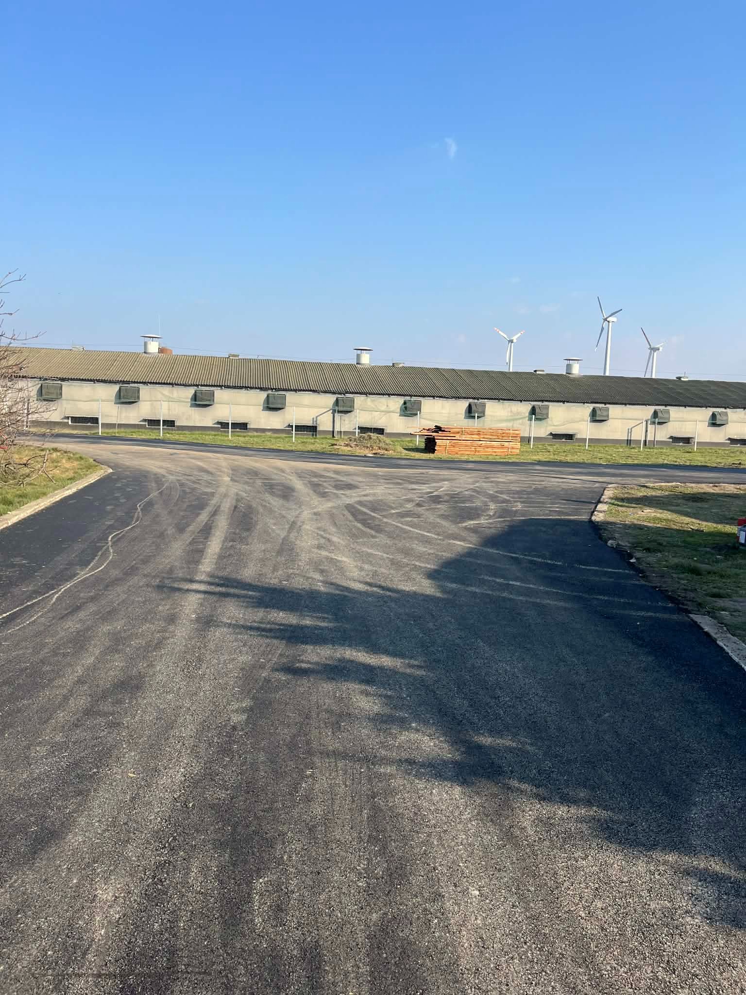 Nowo wyasfaltowana droga dojazdowa z widocznymi śladami opon, prowadząca do budynku gospodarczego z turbinami wiatrowymi w tle, w słoneczny dzień.