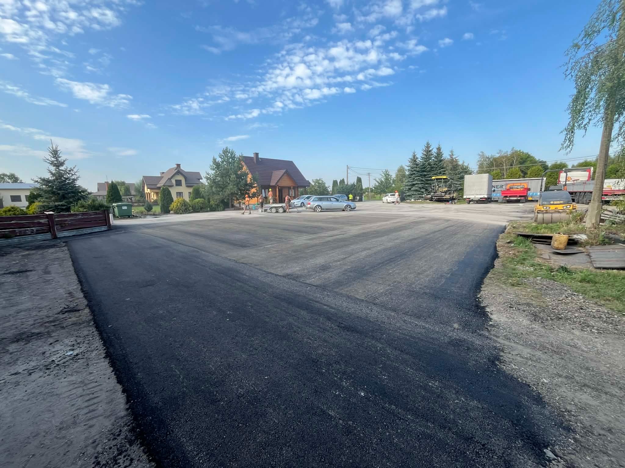 Świeżo położony asfalt na drodze dojazdowej, widoczne maszyny budowlane i ekipa w oddali. Błękitne niebo z chmurami w tle, domy jednorodzinne.