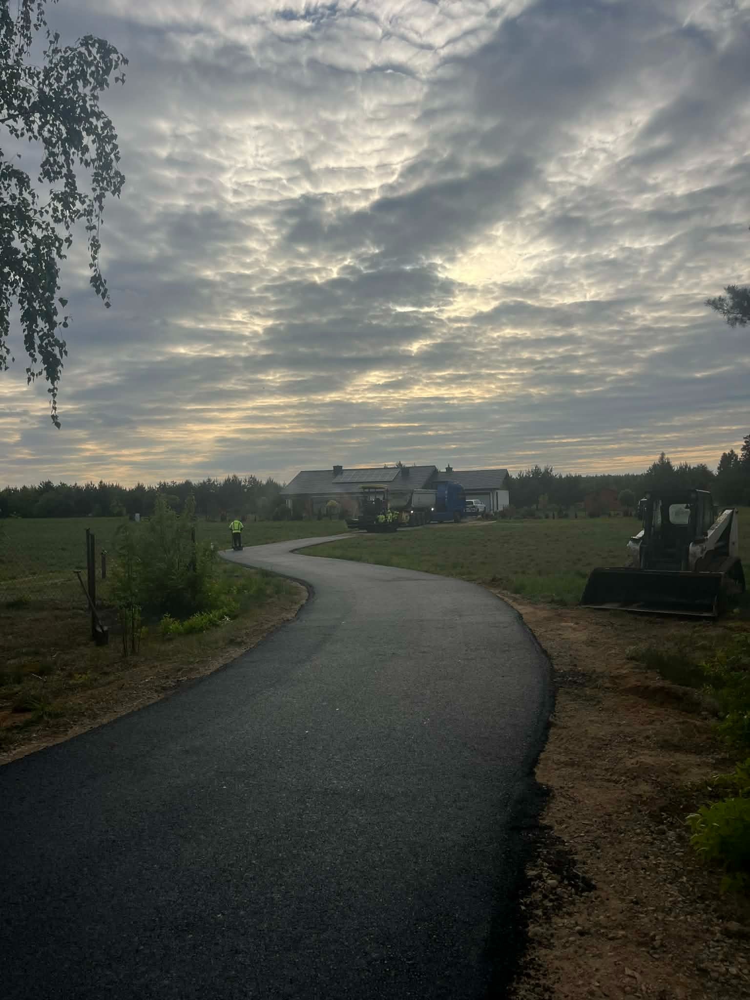 Kręta, asfaltowa droga dojazdowa, świeżo wybudowana, prowadzi w kierunku domu. Na poboczu stoi mała koparka, a w tle widać pracowników w żółtych kamizelkach.