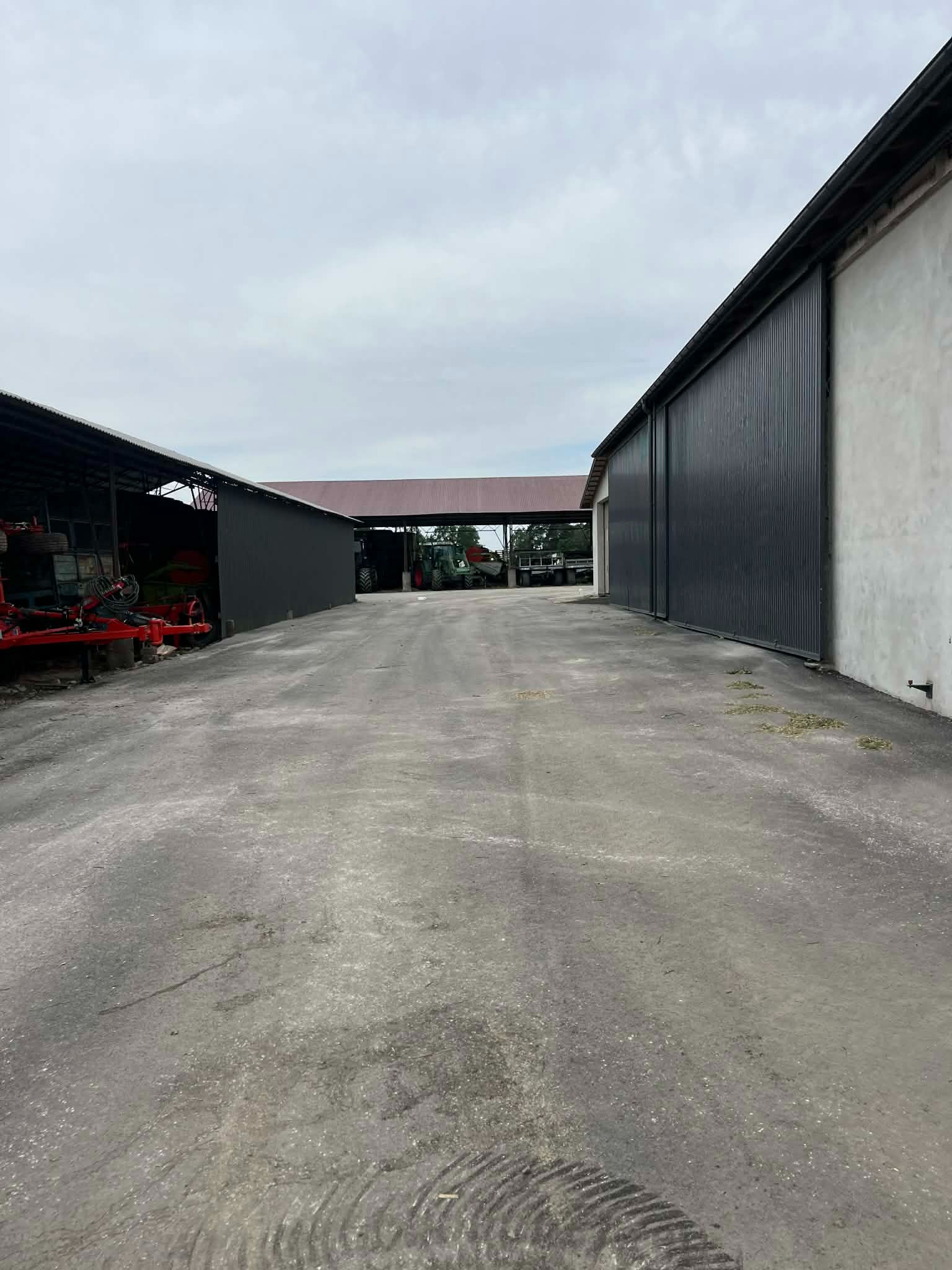 Szeroki, betonowy plac manewrowy z zadaszeniami i maszynami rolniczymi w tle, oświetlony pochmurnym światłem dziennym. Widok w perspektywie.