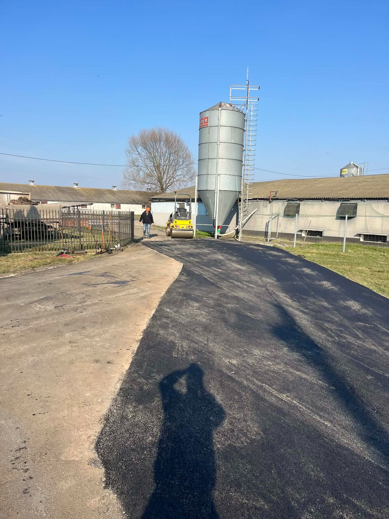 Nowa, czarna nawierzchnia asfaltowa na terenie gospodarstwa, z walcem drogowym i sylwetką pracownika w oddali. W tle budynki gospodarcze i silos.
