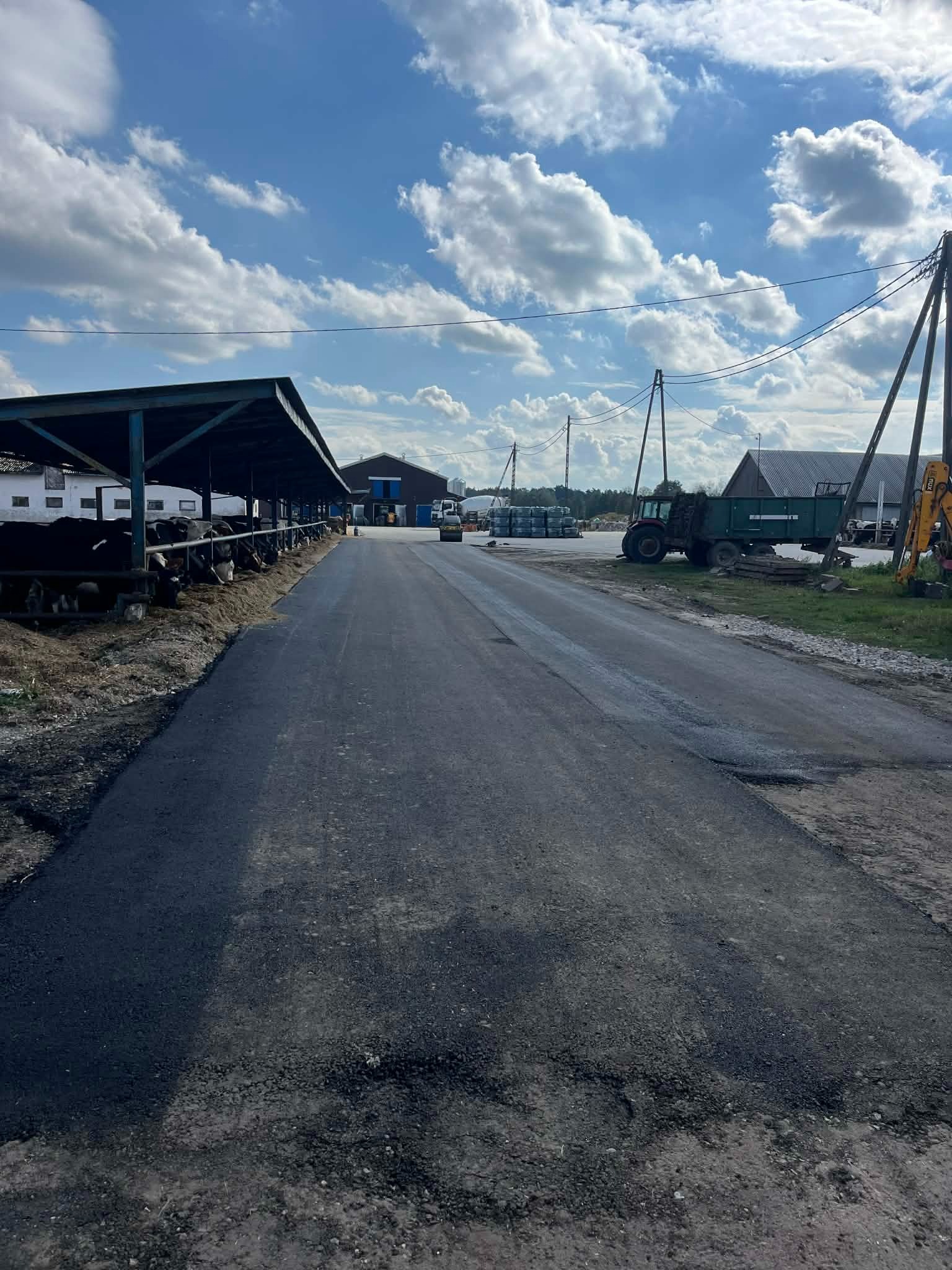 Świeżo wyasfaltowana droga dojazdowa na terenie gospodarstwa rolnego, z widocznymi maszynami budowlanymi i budynkami gospodarczymi w tle, przy słonecznej pogodzie.