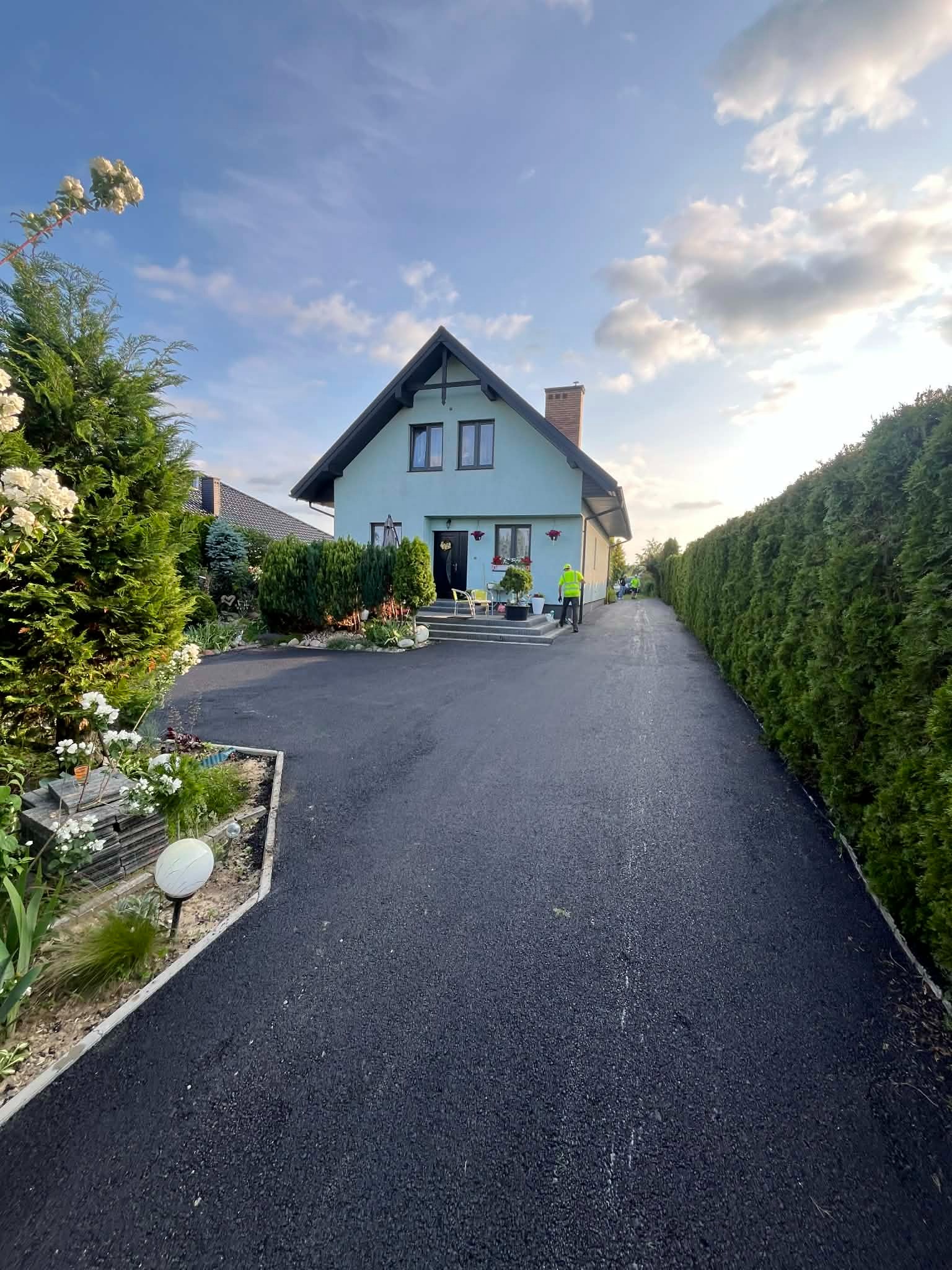 Nowa, asfaltowa droga do domu jednorodzinnego, otoczona zielenią i tujami. Widoczni pracownicy w odblaskowych kamizelkach. Słoneczny dzień, błękitne niebo z chmurami.